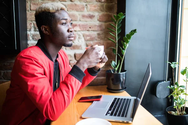 Porträt eines afrikanisch-amerikanischen Mannes in Jacke, der Kaffee trinkt und am Laptop arbeitet — Stockfoto