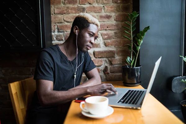 Schöner afroamerikanischer Mann sitzt im Café, hört Musik und schaut auf Laptop — Stockfoto