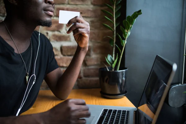 Primer plano de las manos del hombre afroamericano con la tarjeta blanca en blanco para el espacio de copia de adv delante de la computadora portátil y el fondo de la mesa — Foto de Stock