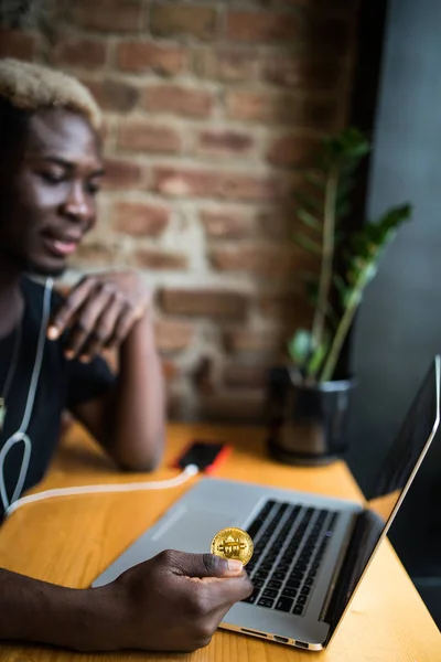 Joven afroamericano hombre sosteniendo bitcoin en la mano delante de la computadora portátil en la cafetería — Foto de Stock