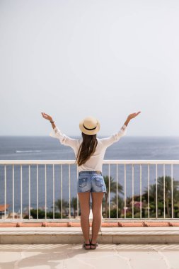 Yaz aylarında genç kadın elbise Hasır Şapkalı bir gökyüzü ve deniz arıyor. Arkadan Görünüm