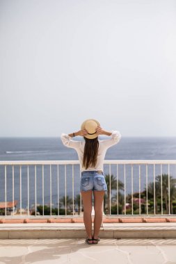 Bir yaz teras üzerinde duran ve Teras okyanusa bakarak hasır şapkalı güzel bir genç kız
