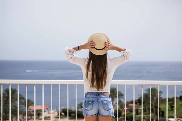 Молодая женщина в летнем платье с соломенной шляпой, смотрящая на небо и море. Вид сзади — стоковое фото