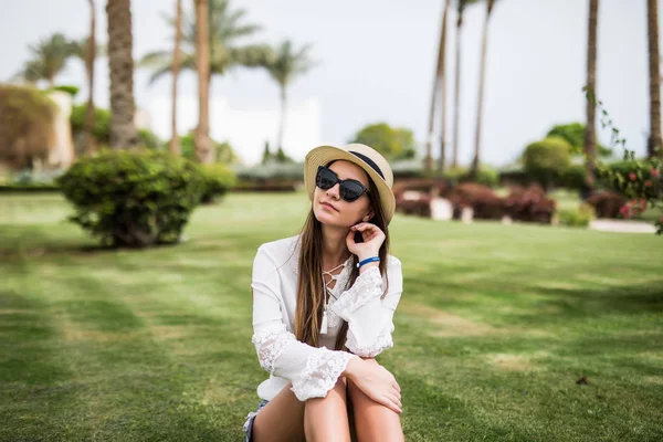 Портрет веселої молодої жінки в літньому капелюсі і сонцезахисних окулярах під пальмою на зеленій траві. Літні канікули — стокове фото