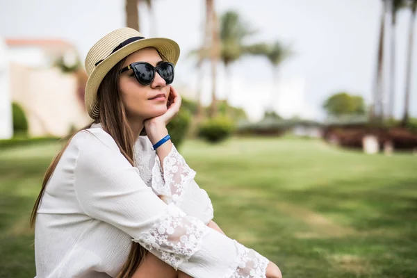 Joyeux jeune femme en chapeau et lunettes de soleil assis sur l'herbe sur fond de palmiers station estivale. vocation estivale — Photo