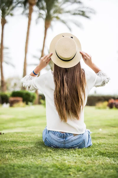 Pohled zezadu mladá kráska žena v klobouku proti slunci seběhla sedí na trávě na pláži pod palmami — Stock fotografie