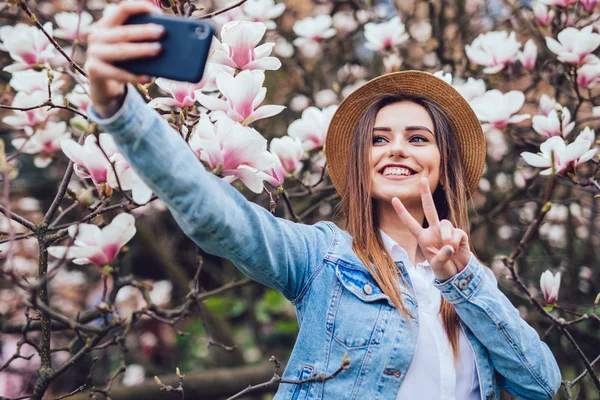 Joven belleza mujer en verano sombrero tomar selfie en el teléfono cerca de flor magnolia árbol en soleado día de primavera — Foto de Stock