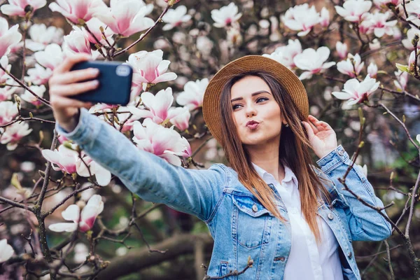 Retrato de primavera de la sensual mujer de belleza en sombrero de paja en el parque del jardín de flores haciendo selfie en el teléfono contra las flores de magnolia — Foto de Stock