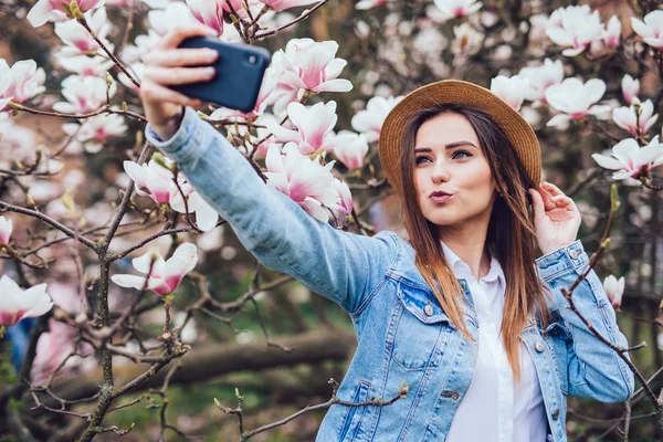 여름 모자에 젊은 아름다움 여자 화창한 봄 날에 꽃 목련 나무 근처 전화 selfie를 받아 — 스톡 사진