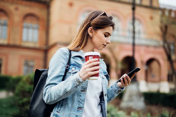 Красивая девушка стоит на улице, печатает по телефону и пьет кофе, чтобы сказать — стоковое фото