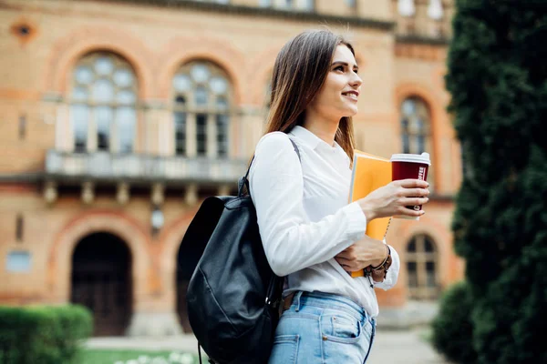 Sorrindo jovem estudante segurando xícara de café, ao ar livre com cadernos, tendo descanso no campus universitário. Conceito de educação, espaço de cópia — Fotografia de Stock