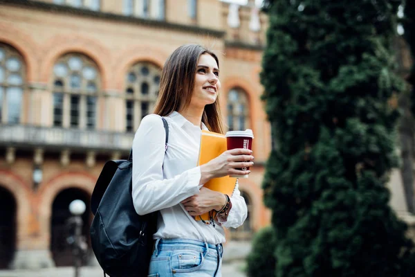 Szczęśliwy uśmiechający się studentka z plecakiem w tło Uniwersytet. Kobieta z skoroszytów i take away kawa o odpoczynku w kampusie w porze obiadowej. Koncepcja edukacji i rozrywki — Zdjęcie stockowe