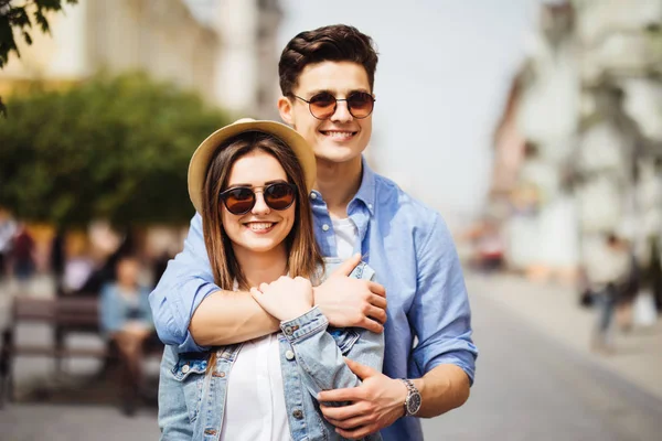 Красива молода пара в сонцезахисних окулярах обіймається, дивлячись на камеру і посміхаючись, стоячи на відкритому повітрі — стокове фото