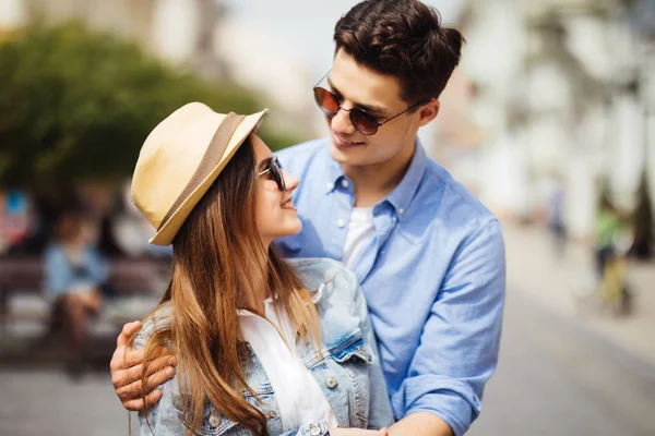 Beau jeune couple en lunettes de soleil étreignant, regardant la caméra et souriant tout en se tenant debout à l'extérieur — Photo