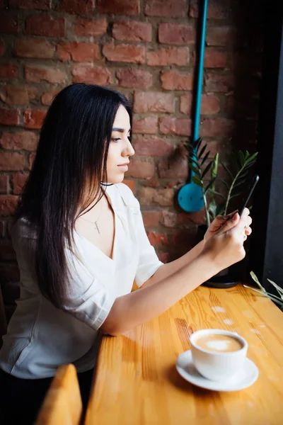 Woman typing write message on smart phone in a modern cafe. Cropped image of young pretty girl sitting at a table with coffee or cappuccino using mobile phone. — Stock Photo, Image