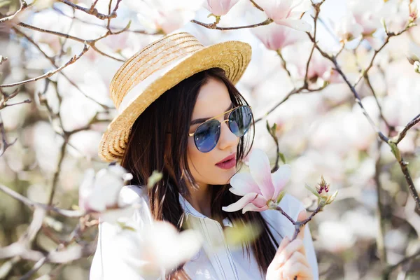 Gyönyörű fiatal nő, kalap és napszemüveg magnólia virág fa körül — Stock Fotó