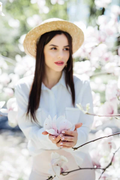 Fiatal szép lány nyári kalap állandó Magnolia virágzó virágok, közel áll a magnolia fa közelében — Stock Fotó