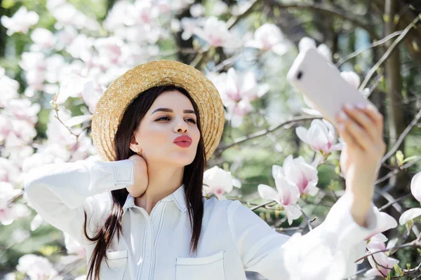 Jeune femme en lunettes de soleil et chapeau de paille prendre selfie au téléphone entouré de magnolia. Saison de printemps — Photo