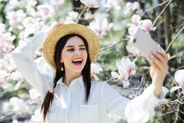 Fiatal nő napszemüveg és szalmakalap fog selfie a telefon körül a magnolia. Tavaszi szezon — Stock Fotó