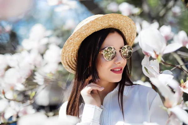 Jonge vrouw in zonnebrillen en stro hoed omringd met magnolia. Lente seizoen — Stockfoto