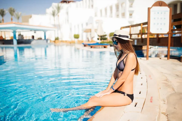 Молодая женщина в солнцезащитных очках и летней шляпе сидит у бассейна. Летнее время — стоковое фото