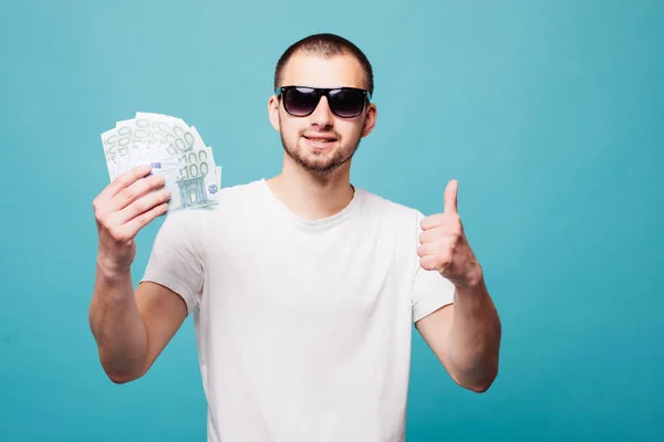 Πορτραίτο ενός άνδρα νέοι καλοκαίρι σε λευκό t-shirt εκμετάλλευση μετρητά χρήματα με τους αντίχειρες επάνω, ενώ χαμογελώντας απομονώνονται σε πράσινο φόντο — Φωτογραφία Αρχείου
