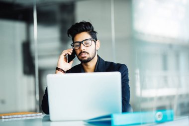 Dizüstü bilgisayarda çalışan ve modern ofisindeki telefonda konuşurken Hintli işadamı