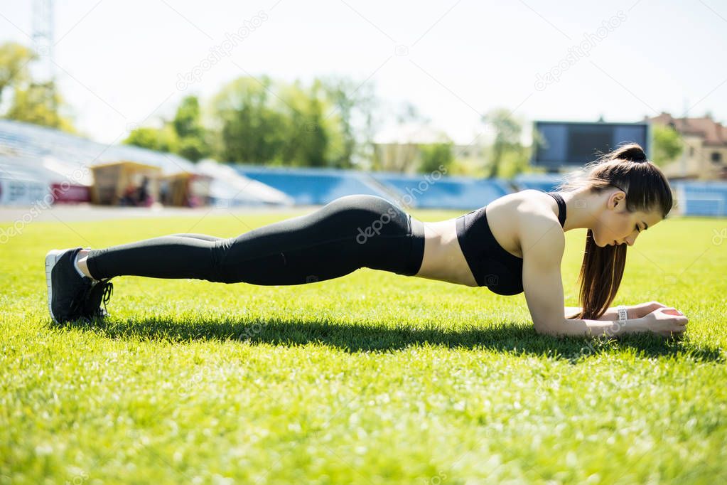 Young happy sportswoman in sportswear making plank exercise on stadium field area outdoors. Healthy lifestyle concept, sport activity.