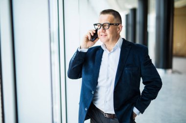 Pozitif kendine güvenen başarılı üst düzey işadamı pencerede ayakta ve Office cep telefonunda konuşurken cityscape düşündüğünü resmi elbiseli