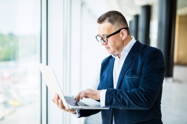 Her zaman size yardımcı olmaya hazır. Onun gözlük ayarlama ve ofiste dizüstü dururken holding resmi kıyafeti içinde neşeli olgun adam