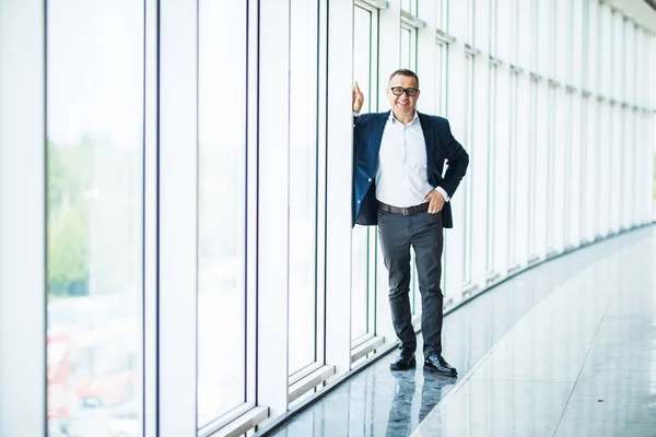 Mature businessman in a corporate suit standing in a large empty office space with modern glass room divider and looking away through large windows optimistically