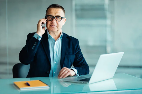 Бізнесмен середнього віку з ноутбуком розмовляє по телефону в сучасному офісі — стокове фото