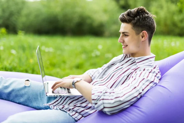 Hombre joven guapo acostado en el sofá inflable lamzac escribiendo en el ordenador portátil mientras descansa en la hierba en el parque —  Fotos de Stock