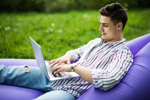 Jeune homme reposant sur un canapé d'air dans le parc. bel homme se détendre sur le canapé gonflable lamzac sur l'herbe avec ordinateur portable — Photo
