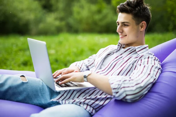 Beau jeune homme couché sur le canapé gonflable lamzac jouer à des jeux sur ordinateur portable tout en se reposant sur l'herbe dans le parc — Photo