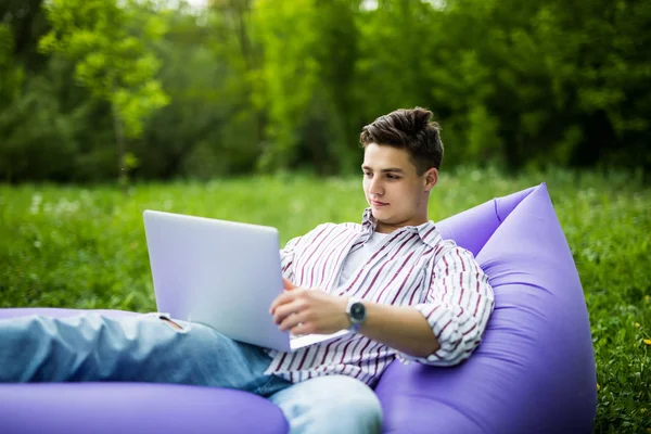 Bello giovane sdraiato sul divano gonfiabile che lavora sul computer portatile mentre riposa sull'erba nel parco — Foto Stock