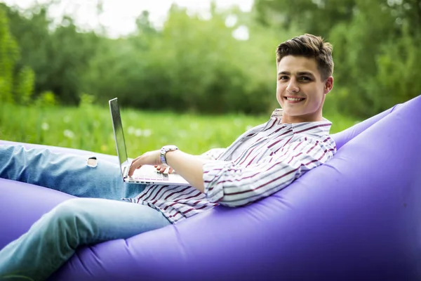 Guapo joven sonrisa hombre acostado en sofá inflable trabajando en el ordenador portátil mientras descansa en la hierba en el parque — Foto de Stock