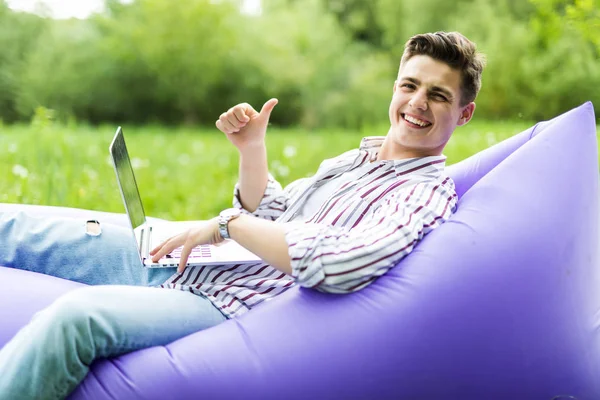 Guapo joven sonrisa hombre acostado en el sofá inflable lamzac trabajando en el ordenador portátil y haciendo gestos pulgares hacia arriba mientras descansa sobre la hierba en el parque —  Fotos de Stock