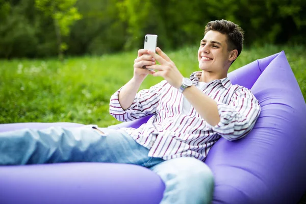 Guapo joven sonrisa hombre acostado en el sofá inflable lamzac navegar en la aplicación en el teléfono mientras descansa en la hierba en el parque — Foto de Stock