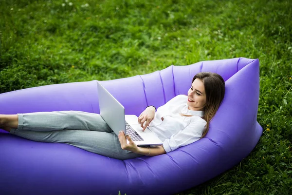 Bella giovane donna sdraiata sul divano gonfiabile lamzac che lavora sul computer portatile mentre riposa sull'erba nel parco — Foto Stock