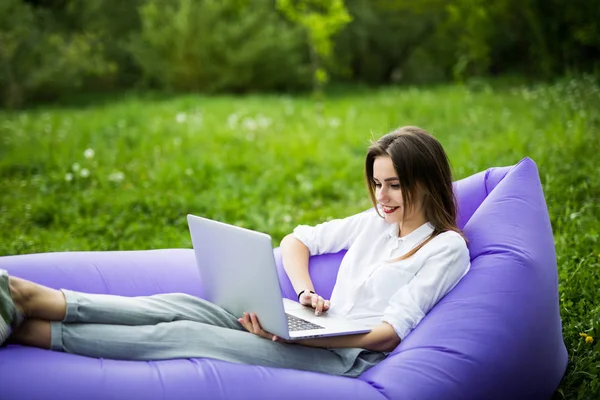 Jolie jeune femme couchée sur un canapé gonflable lamzac utiliser un ordinateur portable tout en se reposant sur l'herbe dans le parc — Photo