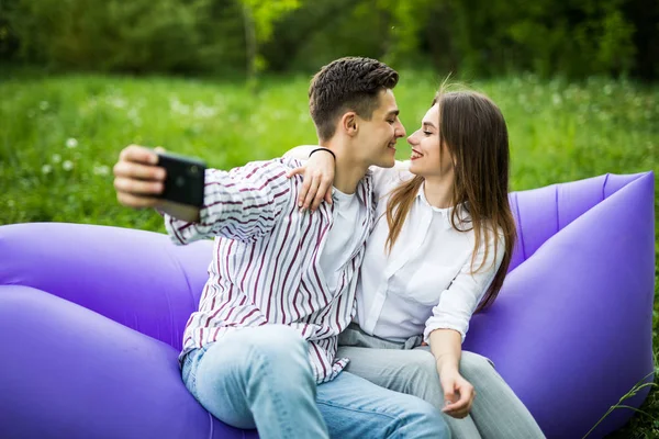 You are my soul. Young couple hugging and kissing while sitting on inflatable sofa lamzac and take selfie on phone resting on grass in park — Stock Photo, Image