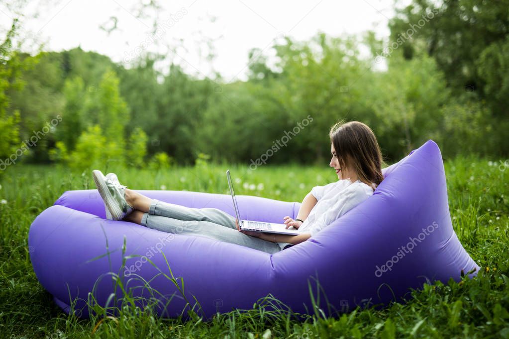 Pretty young woman lying on inflatable sofa lamzac working on laptop while resting on grass in park