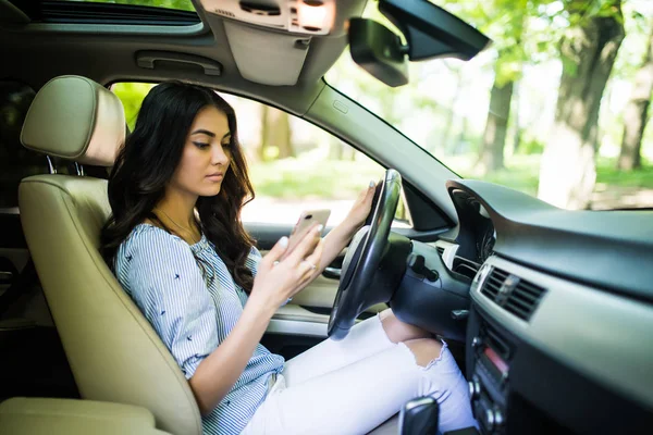 Jovem bonita olhando para seu smartphone enquanto dirige carro na estrada — Fotografia de Stock