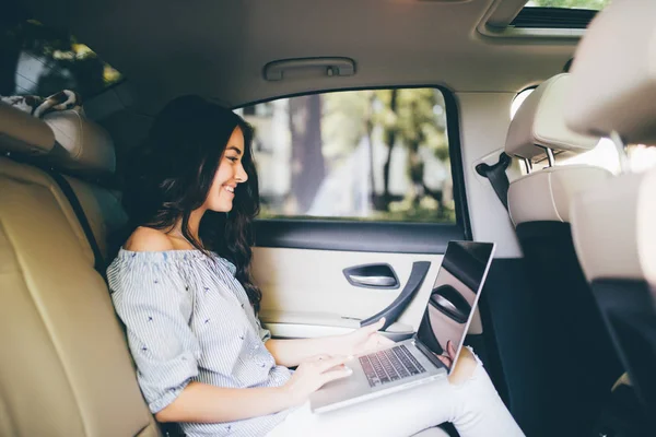 Schöne Geschäftsfrau benutzt einen Laptop und lächelt, während sie auf dem Rücksitz im Auto sitzt — Stockfoto