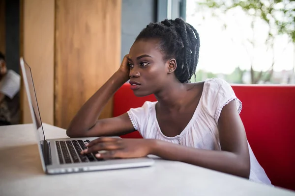 Mujer afroamericana con peinado afro, vestida con capucha blanca casual, sentada en el sofá de la cafetería y esperando a su amiga. Estudiante africano usando laptop y tomando café — Foto de Stock