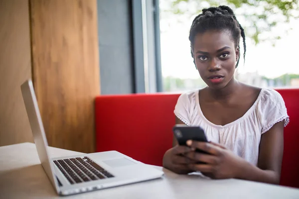 Adorable mujer afroamericana con peinado afro revisando su fuente de noticias o mensajería a través de redes sociales, usando wi-fi gratis en el teléfono móvil, sonriendo, sentada en la cafetería frente a la computadora portátil — Foto de Stock