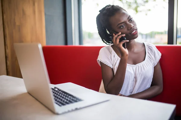 Retrato de una chica afroamericana sentada en la mesa de la cafetería y trabajando en su computadora portátil hablando en su teléfono celular mientras cuidadosamente mira a un lado — Foto de Stock