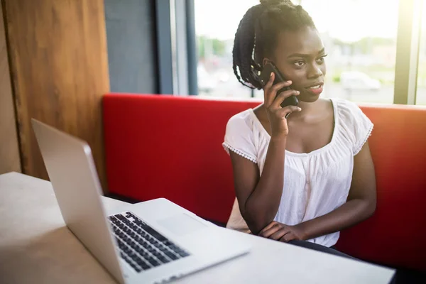 Concepto de tecnología y comunicación. Exitosa mujer de negocios afroamericana con peinado afro que hace llamadas telefónicas con mirada seria y segura mientras trabaja en un café en una computadora portátil — Foto de Stock