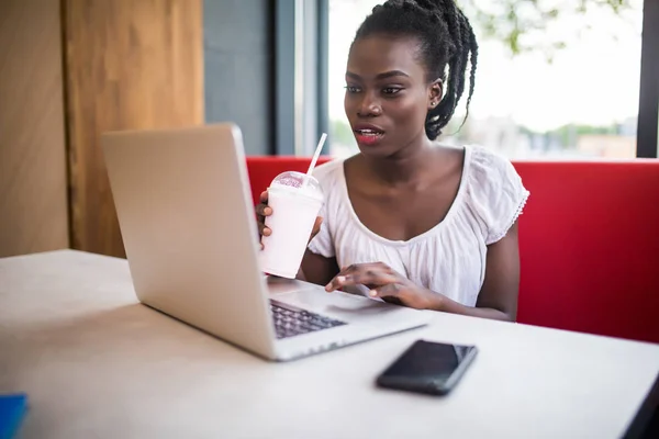 Retrato de una estudiante afroamericana escribiendo respuestas a amigos mientras trabaja en una computadora portátil con conexión inalámbrica a Internet bebiendo batido en la cafetería — Foto de Stock